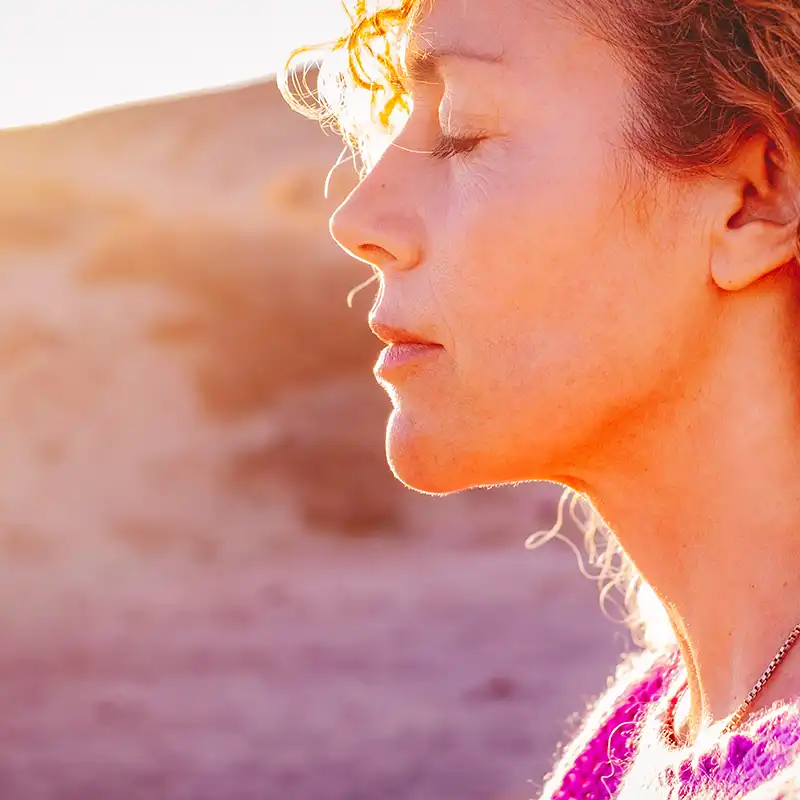 Nahaufnahme eines Sonnenuntergangsporträts einer attraktiven Frau mit geschlossenen Augen und der Sonne im Gegenlicht. Träumendes und genussvolles Gefühl, Lifestyle-Emotion. Gelassene Frau im Freien mit lockigem Haar.